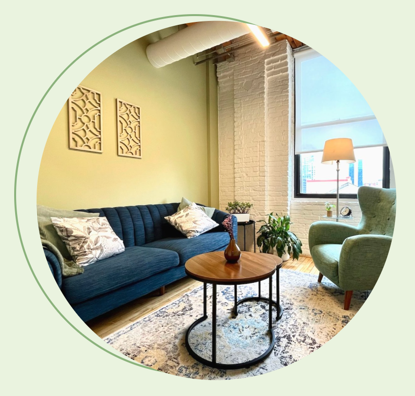 Chicago in-person session office — a warm, welcoming therapy room with a blue sofa, green armchair, round wooden coffee table, and exposed brick wall with natural light